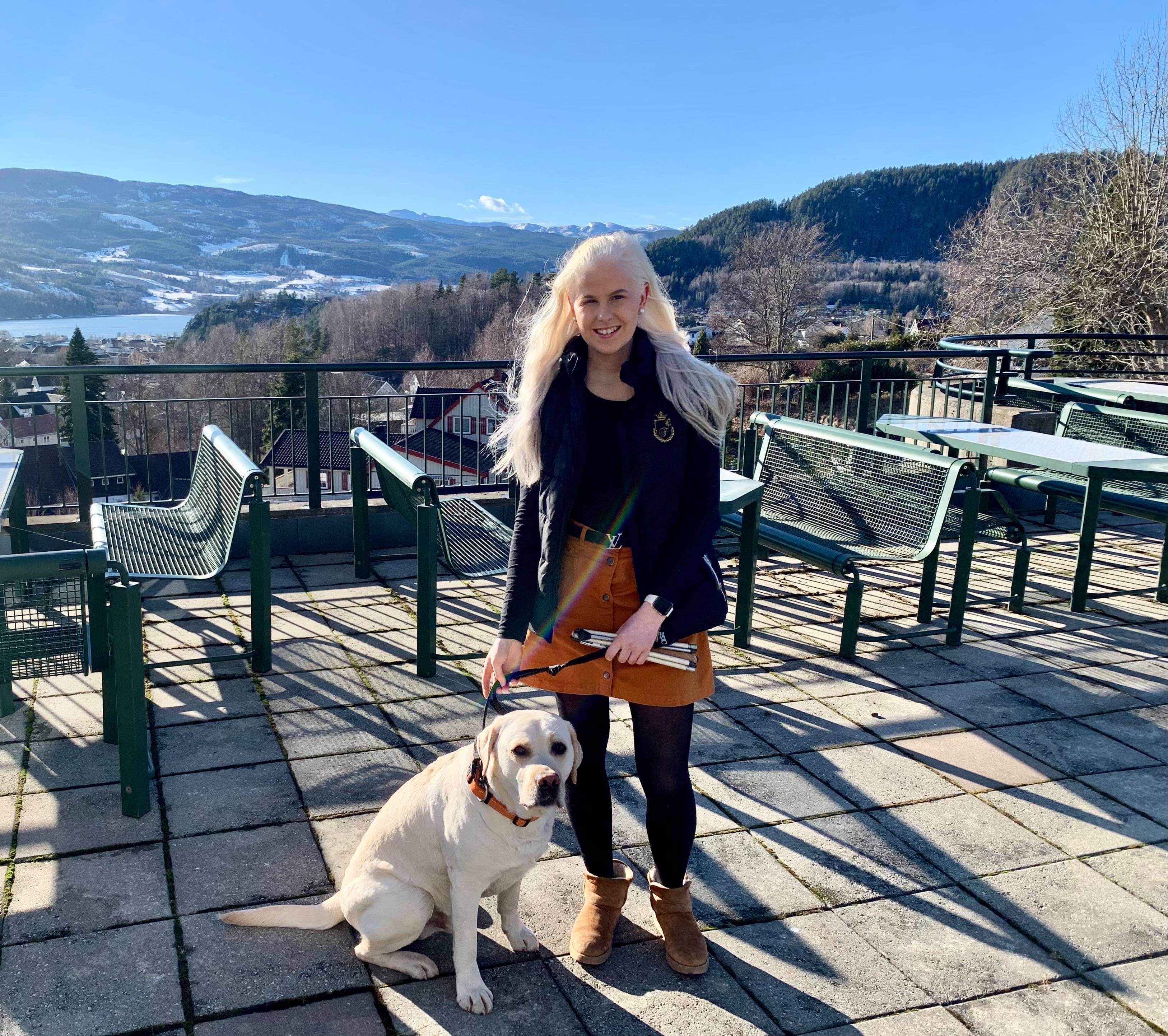 Bilete syner Silje Solvang på tur med hunden Aro. Ho står ute på ein terrasse, medan den lyse labradoren sit ved føtene hennar. Silje smiler mot kameraet, og bak henne skiner sola mot blå himmel.