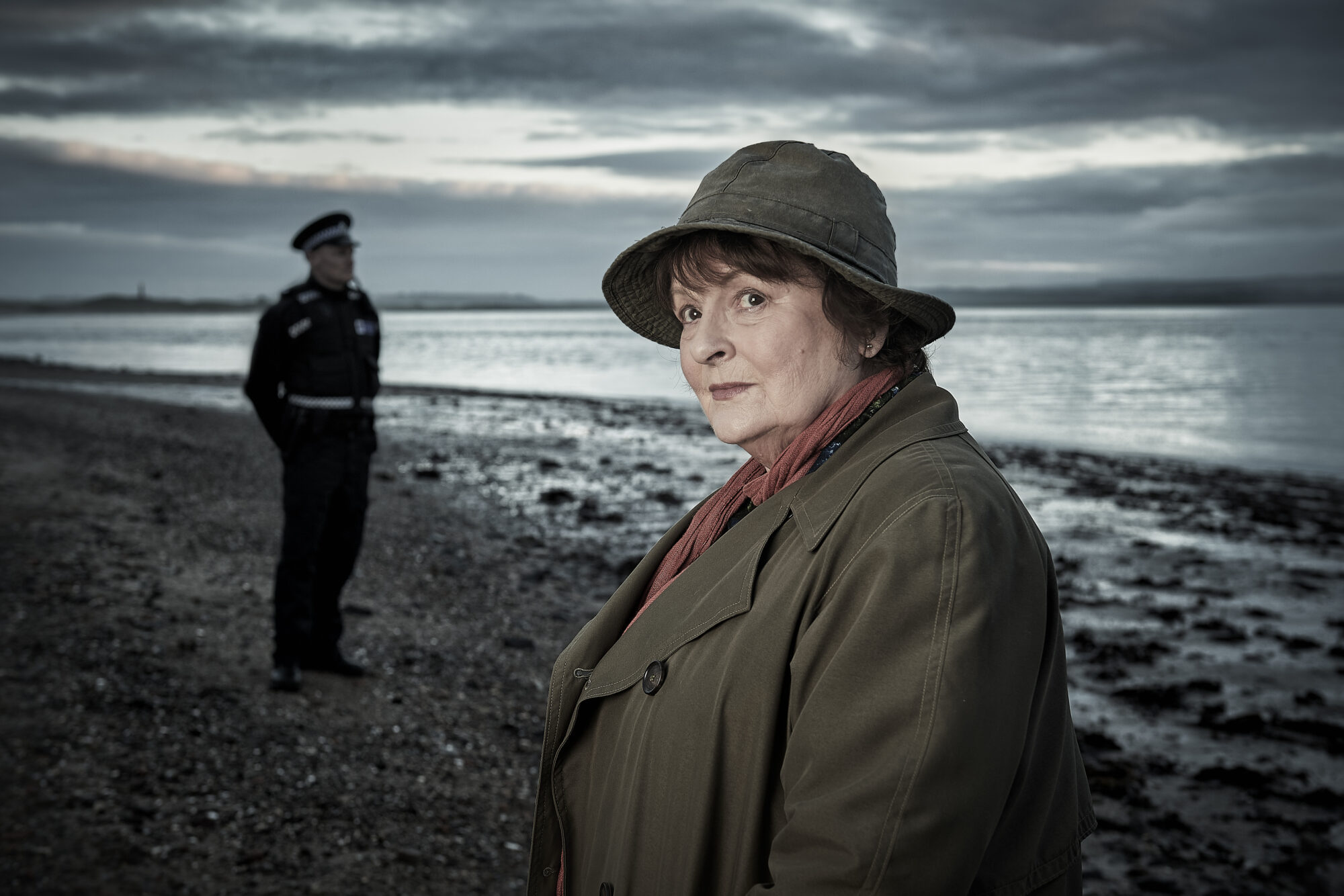 Etterforskar Vera står på ei strand i sitt karakteristiske antrekk, med hatt og frakk. I bakgrunnen står ein politimann i uniform.