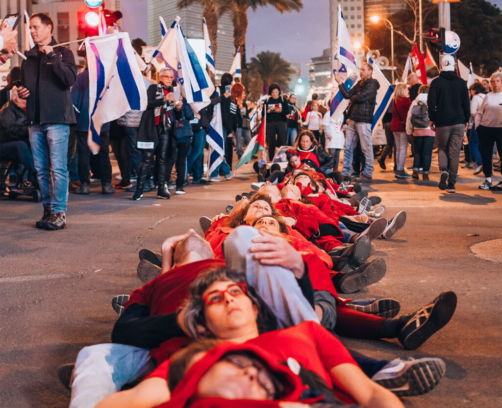 Folkemengde med israelske flagge, og ei gruppe menneske i raudt som har lagt seg ned på bakken.