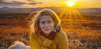 Kvinne med blondt hår og friluftsklær ligger på en skinfell i solnedgang