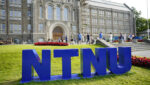 Dei store blå bokstavane NTNU står oppstilt framfor eit grått mursteinsbygg.