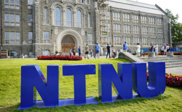 Dei store blå bokstavane NTNU står oppstilt framfor eit grått mursteinsbygg.