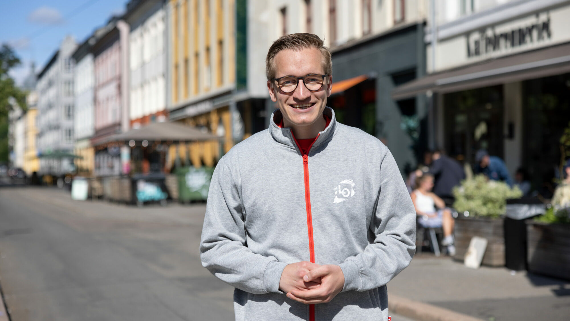 Ung mann med briller og grå jakke smiler, medan han står i ei gate