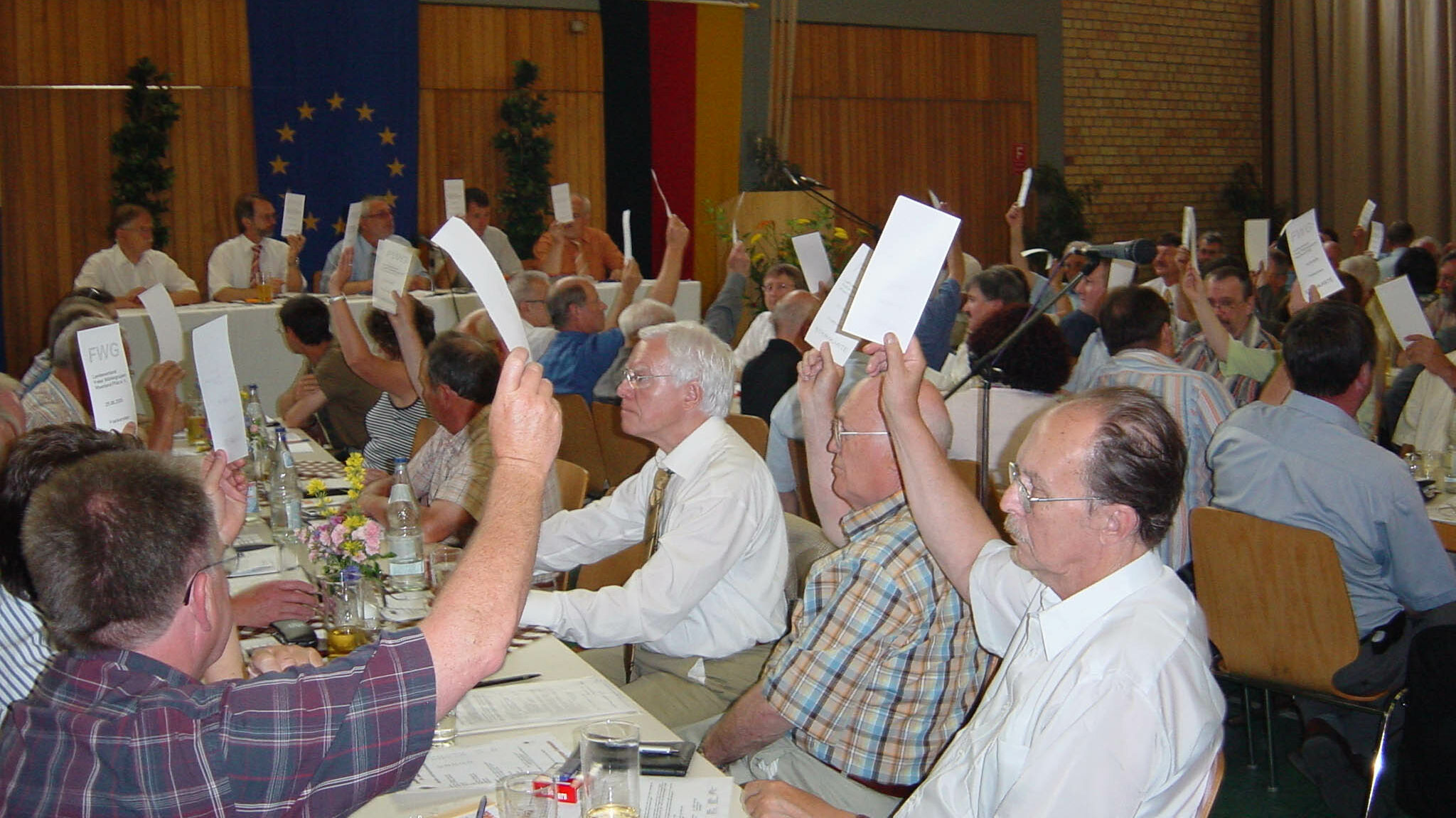 Old men, and a very few women, are sitting together in a large room.  They held white papers and participated in the vote.