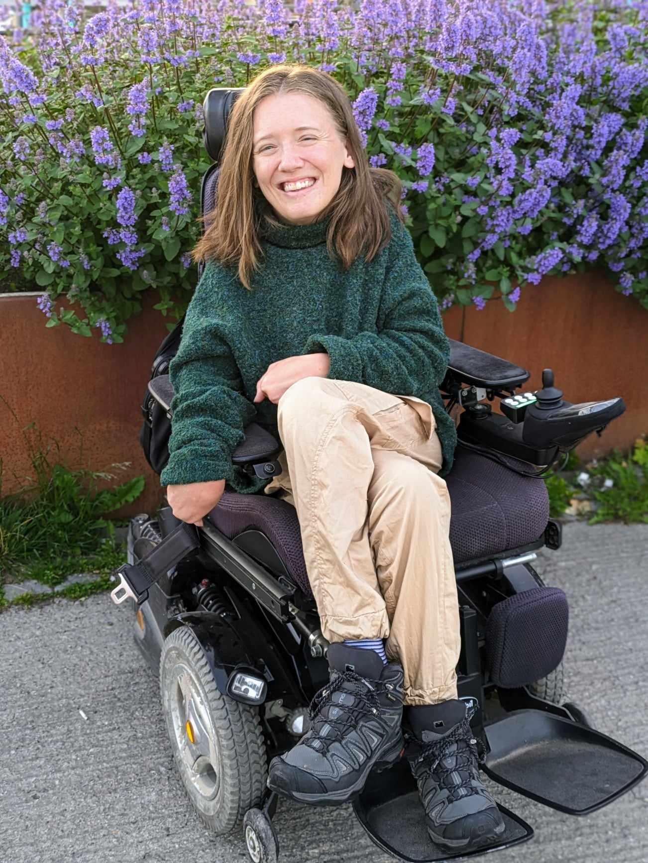 Ung kvinne i rullestol smiler mot kameraet, med lilla blomar i bakgrunnen.