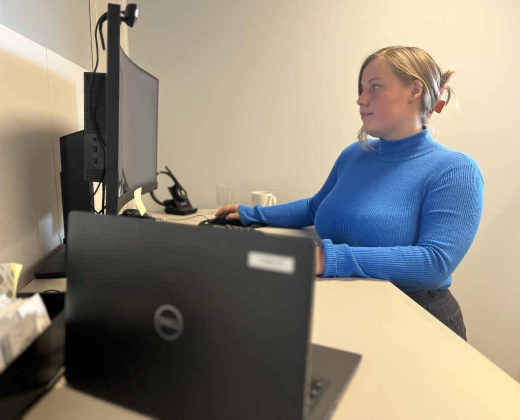 Blond kvinne i blå genser ved datamaskin.