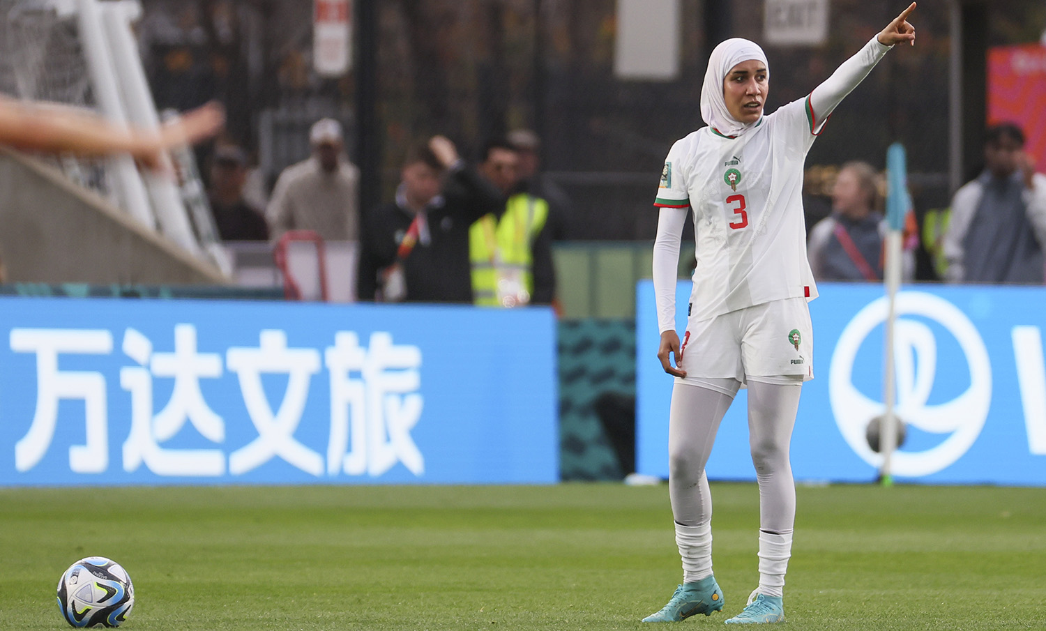 Kvinne på fotballbane i kvit fotballdrakt med langerma skjorte, heildekkande strømper og hijab peikar medan ho har ballen framfor seg, klar til avspark.