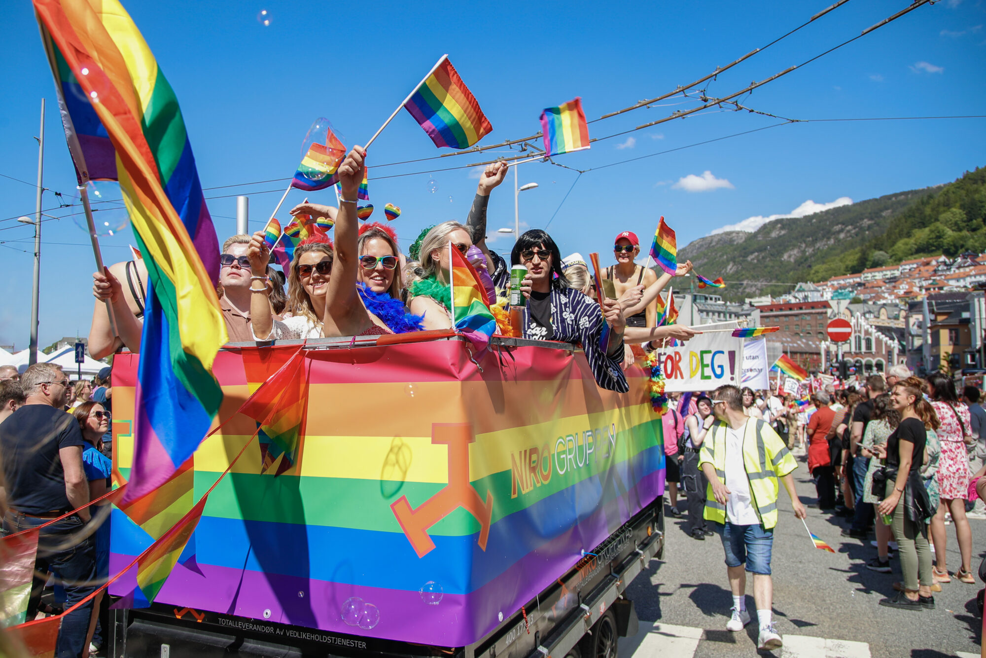 Pride-flåte i strålande sol i Bergen. Flåten er kledd i regnbogefargane og dei ombord viftar med regnbogeflagg.