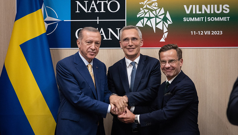 Tre middelaldrene menn står framfor ein Nato logo og forsøker. å ha eit felles handtrykk.