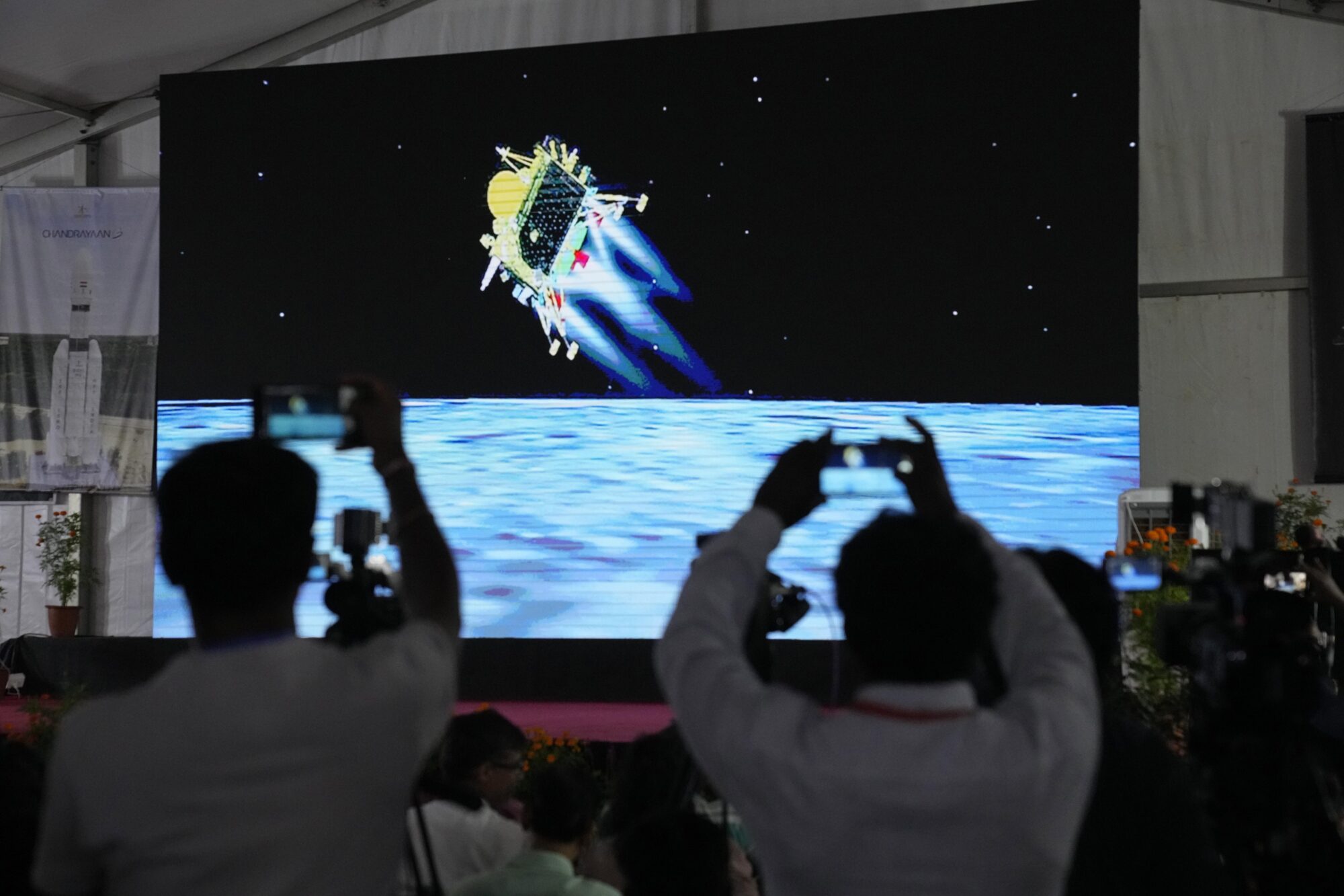 Skjerm viser romfartøy på veg til å lande på månen, medan folk filmar det som skjer.