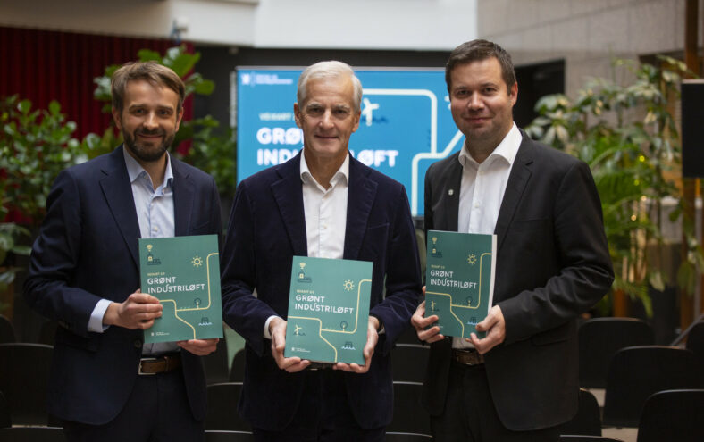 Tre menn i dress viser stolt fram ei grøn bok som dei alle held i hendene sine.