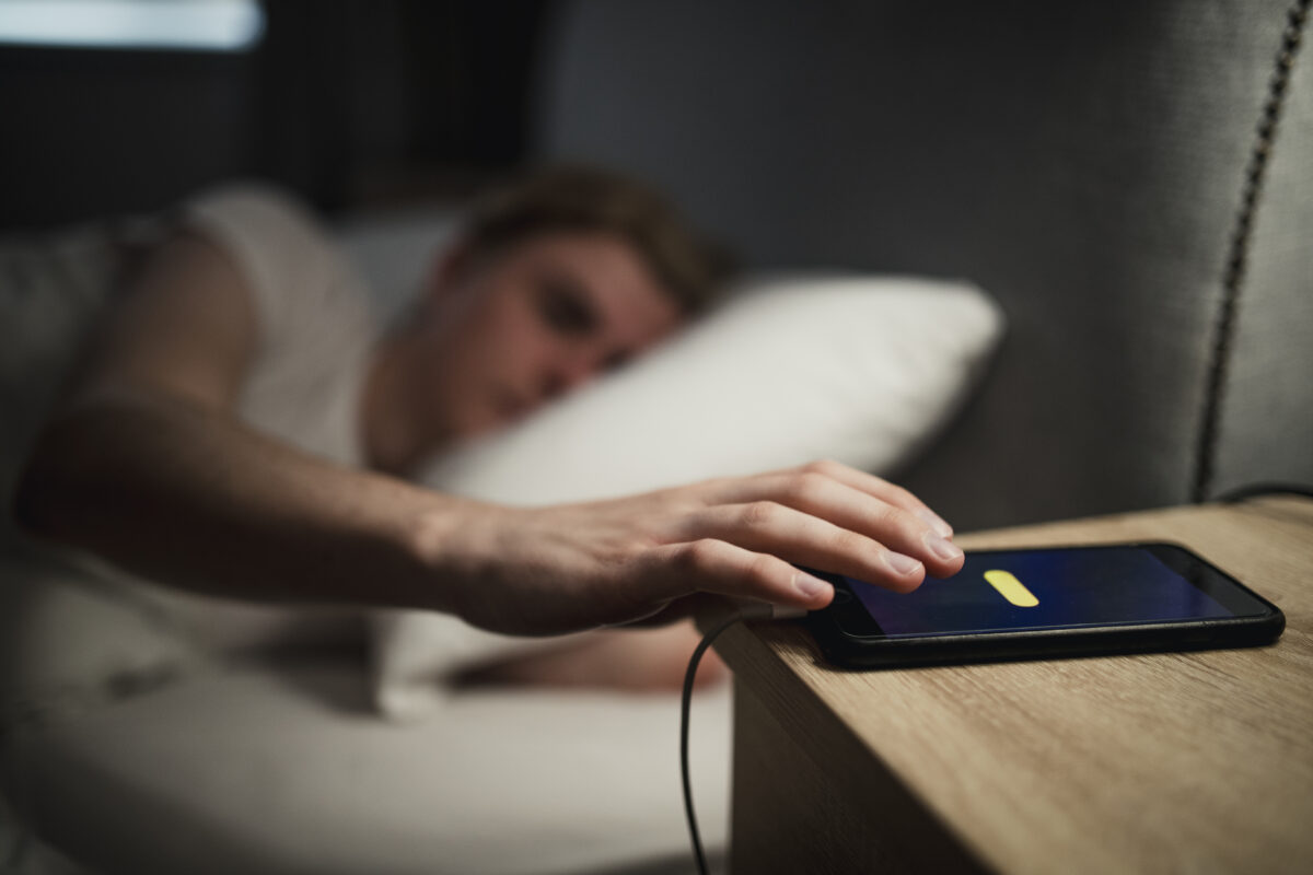Person ligg i senga og strekker arma mot telefonen på nattbordet for å slumre alarmen.