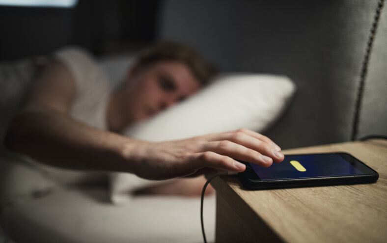 Person ligg i senga og strekker arma mot telefonen på nattbordet for å slumre alarmen.