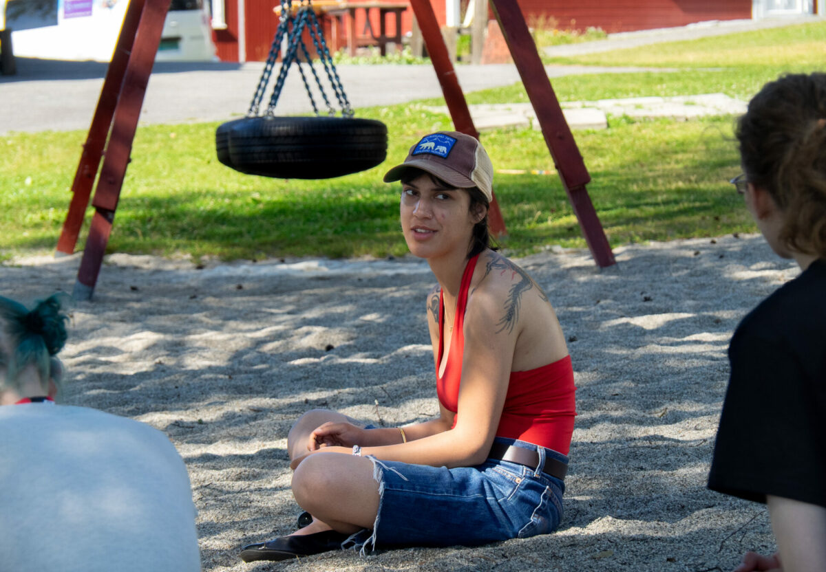 Leiaren for RU, Amrit Kaur, sit på sand. Ho har på ein blå jeans-shorts, og ein raud singlet.