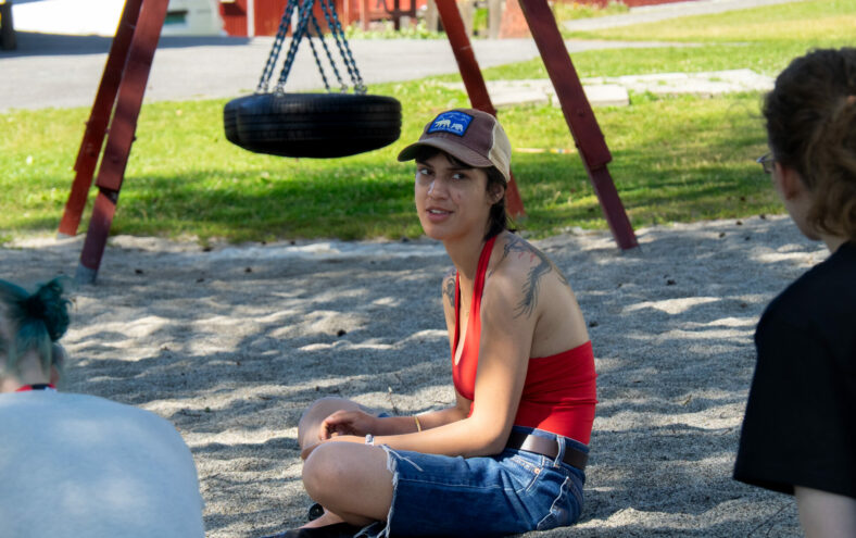 Leiaren for RU, Amrit Kaur, sit på sand. Ho har på ein blå jeans-shorts, og ein raud singlet.
